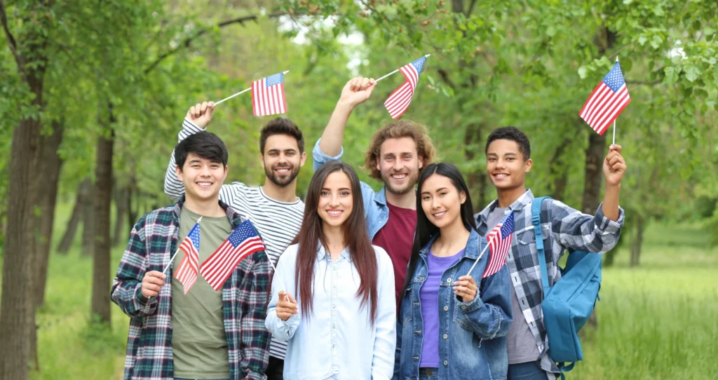 A diverse group of smiling young adults standing outdoors in a park, holding small American flags and posing together.