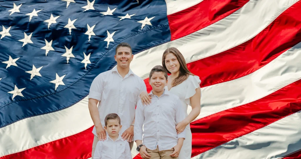 A smiling family of four dressed in white stands together in front of a large American flag backdrop.