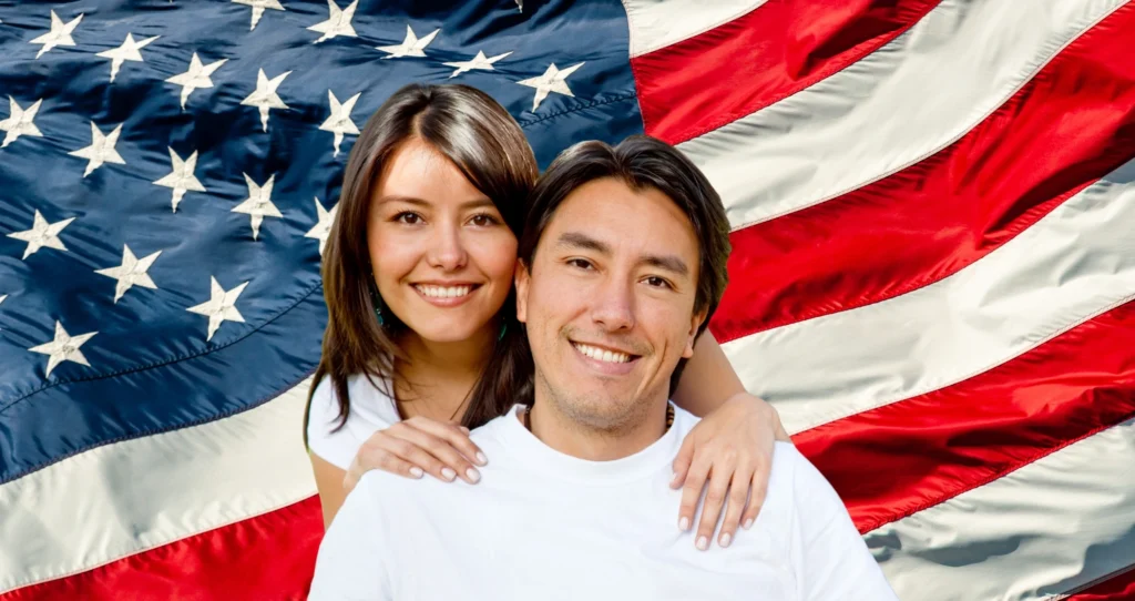 A smiling couple wearing white poses together in front of a large American flag backdrop.
