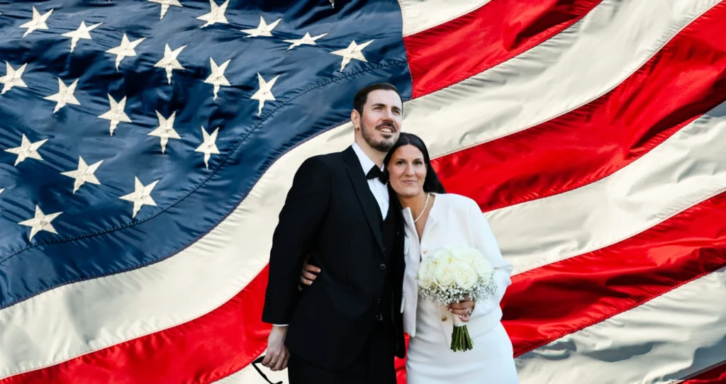 “Couple in wedding attire standing together in front of the U.S. flag.”