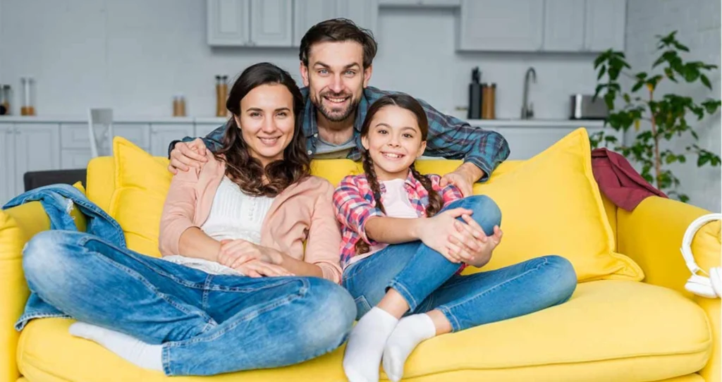 A smiling family of three—two adults and one child—sitting together on a yellow couch in a bright living room.