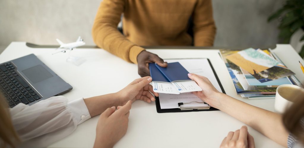People sitting at a desk exchanging travel documents and boarding passes, with a passport, laptop, miniature airplane, and travel brochures visible.